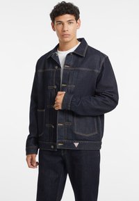 Dark denim jacket with contrasting yellow stitching, button closures, two chest pockets, and a triangle logo detail at the hem.