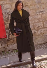 Palton lung negru cu guler lat și cordon în talie, accessorii cu cizme maro închis până la genunchi și o geantă de mână pliabilă maro.