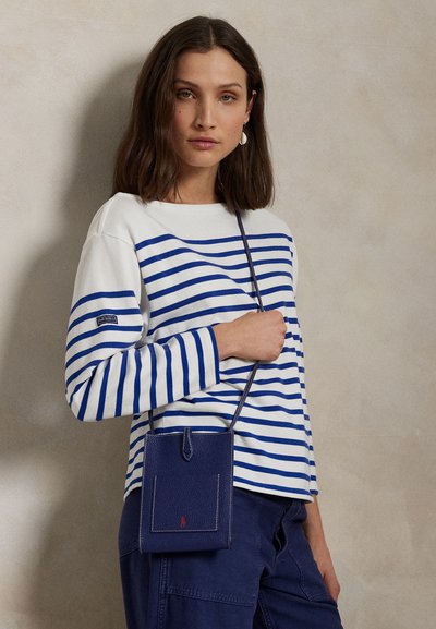 Donna con capelli castani fino alle spalle indossa una camicia a righe blu e bianche a maniche lunghe, pantaloni blu navy e tiene una piccola borsa a tracolla blu navy.