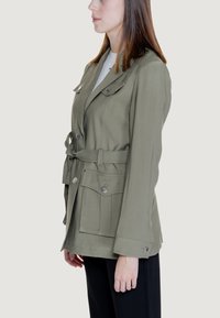 Giacca verde oliva realizzata in tessuto testurizzato, con vita cintrata, ampie tasche frontali e dettagli in metallo argentato.