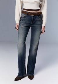 Jeans i denim med rak benpassform, mörkblå tvätt, subtil blekning och bälteshällor; kombinerat med en ljuskrämfärgad tröja och brunt bälte.