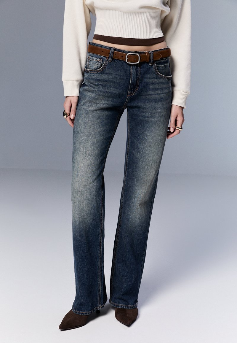 Jeans i denim med rak benpassform, mörkblå tvätt, subtil blekning och bälteshällor; kombinerat med en ljuskrämfärgad tröja och brunt bälte.