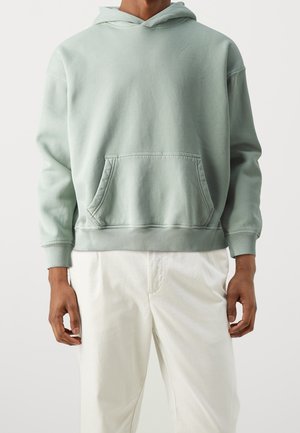 Person wearing a light green hooded sweatshirt with front pocket and white pants, standing against a plain light background.