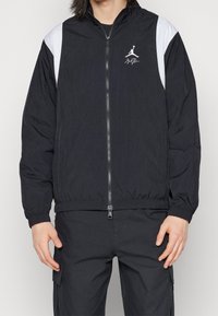 Černá lehká bunda s bílými akcenty, celým zipem, elastickými manžetami a pasem. Na levé straně hrudi je logo.