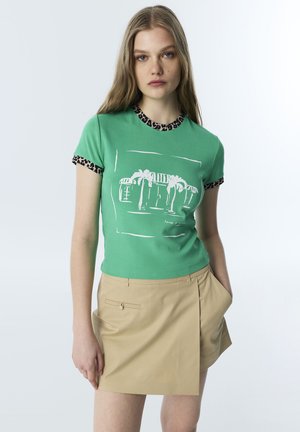 Jonge vrouw die een groen T-shirt met palmboomprint en luipaardbies draagt, gecombineerd met een beige wikkel minirok, staand tegen een effen achtergrond.