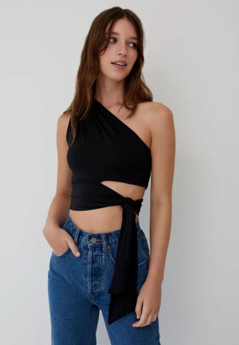 Jonge vrouw die een zwart cropped top met één schouder en zijknoop draagt, gecombineerd met een high-waisted blauwe jeans, staand tegen een effen muur.