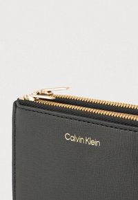 Μαύρο δερμάτινο πορτοφόλι με φερμουάρ σε χρυσαφί τόνο, με το ανάγλυφο όνομα της μάρκας «Calvin Klein» στο μπροστινό μέρος. Επιφάνεια με υφή.