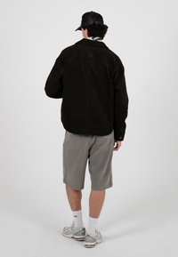 Mann trägt schwarze Cordjacke, graue Shorts, weiße Socken, Turnschuhe und schwarze Baseballkappe, steht mit dem Rücken zur Kamera vor weißem Hintergrund.