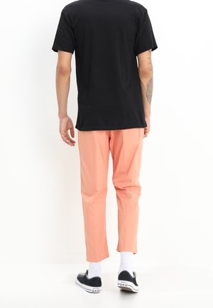 Pakaian santai yang menampilkan kaos hitam lengan pendek dan celana tapered berwarna pink muda. Celana tersebut memiliki tekstur yang halus dan dipadukan dengan sepatu sneakers hitam.