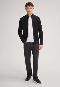 Zwart geribd vest met rits, gedragen over een wit T-shirt, gecombineerd met donkere jeans en zwarte sneakers. De textuur is duidelijk geweven.