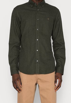 Mann trägt ein geknöpftes dunkelgrünes Hemd mit langen Ärmeln und Kragen und beige Hose, steht vor einem einfarbigen Hintergrund.