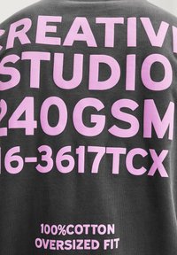 Sivi pamučni dukserica s prevelikim krojem, koja sadrži velike ružičaste natpise: "KREATIVNI STUDIJA," "240GSM," i "100% PAMUK" koji su istaknuti.