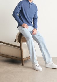 Niebieska koszulka polo z długim rękawem i kołnierzykiem, w zestawieniu z jasnoniebieskimi spodniami prostymi i białymi trampkami, siedząca na drewnianym krześle z poduszką.