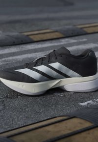 Chaussure de course Adidas noire et blanche avec une grande semelle étiquetée 36, reposant sur une route asphaltée avec des dos d’âne en arrière-plan.