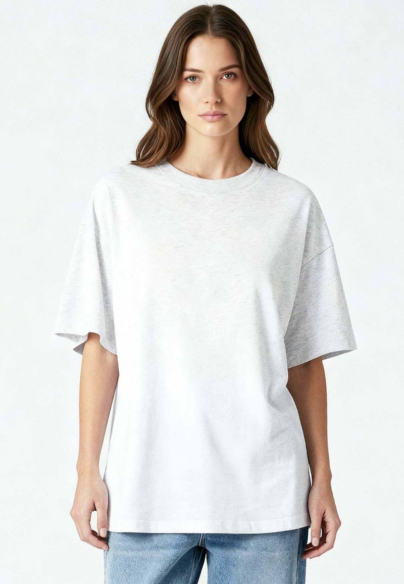 Jeune femme aux longs cheveux bruns portant un t-shirt ample gris clair et un jean bleu, debout devant un fond blanc uni.