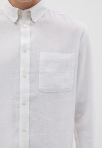 Camicia bianca in lino con colletto button-down, caratterizzata da una singola tasca sul petto e da una sottile texture su tutto il tessuto.