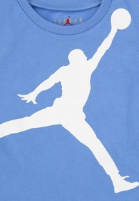 Světle modré bavlněné tričko s velkou bílou siluetou osoby, která skáče a sahá po basketbalovém míči, s kulatým výstřihem.