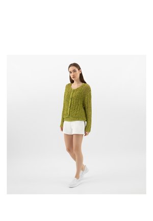 Junge Frau trägt eine olivgrüne Strickjacke, weiße Shorts und weiße Slip-on-Schuhe und steht vor einem schlichten hellen Hintergrund.
