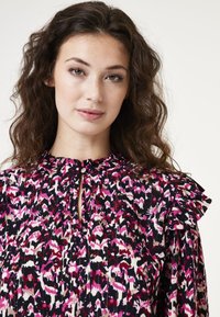 Bloes met bloemenprint en geplooide schouderdetails, met een keyhole-halslijn. Kleuren zijn zwart, roze, crèmekleurig en bordeaux op een geweven stof.