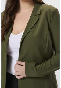Femme portant un blazer vert olive sur un haut blanc et un pantalon noir, tenant le blazer fermé d'une main près du bouton.