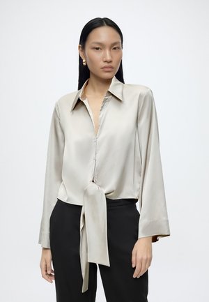 Femme aux longs cheveux noirs portant un chemisier beige en satin boutonné noué à la taille et un pantalon noir, debout sur un fond blanc.