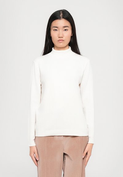 Jeune femme avec de longs cheveux noirs portant un pull à col roulé blanc et un pantalon en velours côtelé beige, se tenant devant un fond clair.