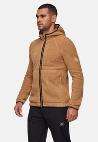 Giacca in pile color sabbia con zip, cappuccio e rifiniture nere. Presenta una tasca frontale e un dettaglio del logo sulla manica. Tessuto morbido.