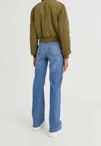 Person seen from behind wearing an olive green bomber jacket, high-waisted blue jeans with back pockets, and white sneakers.