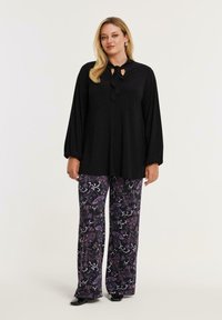 Kvinde stående iført en sort langærmet bluse med en halsbind og løse bukser med blomsterprint i lilla og sort.