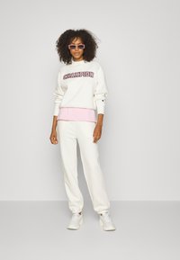 Hvit genser med "Champion"-logo, lagd over topp i rosa; kombinert med hvite joggebukser. Tilbehør inkluderer rosa solbriller og hvite sneakers.