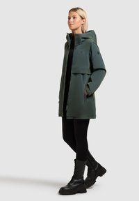 Kobieta ubrana w ciemnozieloną parka z kapturem i kieszeniami, czarne legginsy oraz czarne, sznurowane buty wojskowe, stojąca bokiem na jasnym tle.