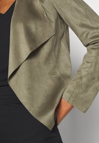 Giacca in suede sintetico verde oliva con fronte aperto, maniche a tre quarti e colletto drappeggiato. Texture liscia e dettagli di design a strati.