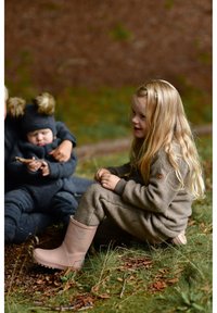 Laps helepruunist villast kombinesoonist koos nahast sildiga, seljas roosad kummisaapad. Pikad lainelised heledad juuksed; istub murul koos lehtedega.