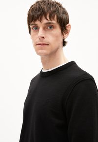 Schwarzer Strickpullover mit Rundhalsausschnitt, ausgestattet mit strukturierten Ärmeln und einem dezenten Logodetail auf der Brust. Weicher Stoff.
