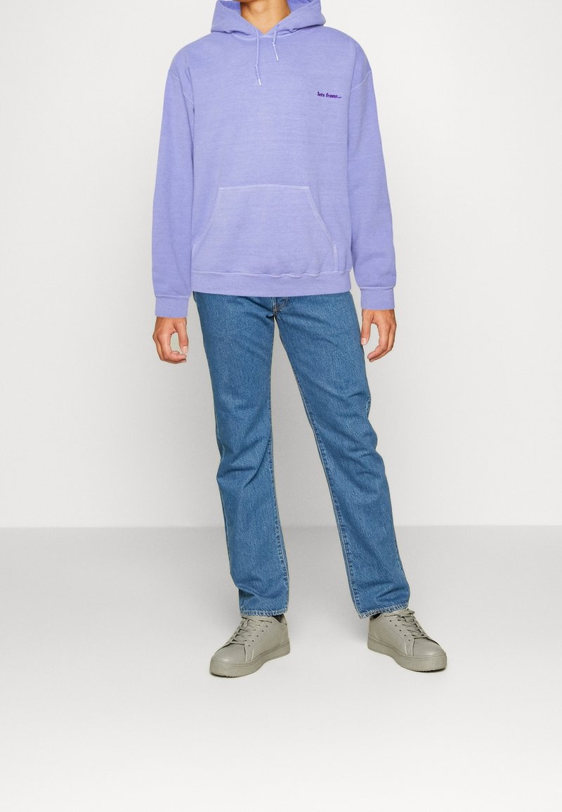 Felpa lavanda con tasca frontale e testo ricamato, abbinata a jeans azzurri chiari e sneaker grigie. Design semplice e vestibilità casual.