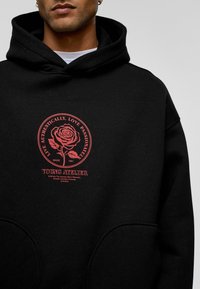 Zwarte hoodie met een grote rode graphic van een roos en tekst. Gemaakt van zachte stof, met een voorzak en een capuchon met trekkoord.