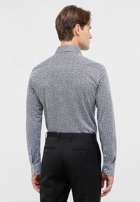 Langarmshirt in blauw-witte bloemenpatroon, gemaakt van zachte stof. Heeft een spreadkraag en knoopmanchetten. Gecombineerd met zwarte broek.
