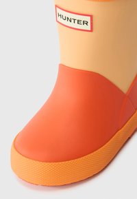 Oranžni gumijasti dežni škorenj z gladko teksturo, ki ima kontrastno svetlejši zgornji del in teksturiran oranžen podplat. Na strani je oznaka z logotipom.