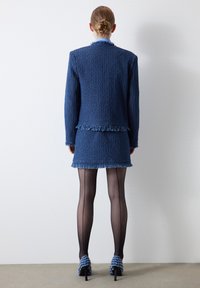 Costume en tweed bleu avec un ourlet frangé, comprenant une veste ajustée et une mini-jupe. Mannequin portant des collants noirs transparents et des talons à motifs.