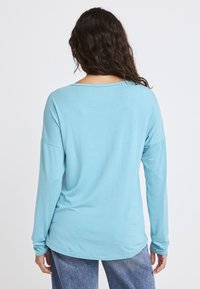 Jasnoniebieska koszula z długim rękawem o luźnym kroju, zaokrąglonym dekolcie i miękkiej fakturze. Posiada opadłe ramiona i zaokrąglony dół.