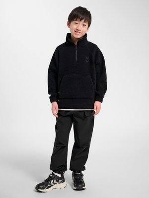 Junger Junge steht, trägt schwarzen Fleecepullover, schwarze Hose und schwarze Sneaker mit weißen Sohlen, lächelt in die Kamera vor einfachem Hintergrund.