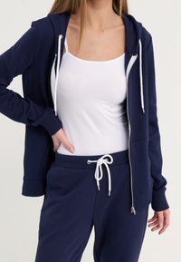 Sweat à capuche zippé bleu marine avec cordons blancs, assorti à un pantalon de jogging. Tissu en coton doux, coupe décontractée, avec poches latérales et taille réglable.