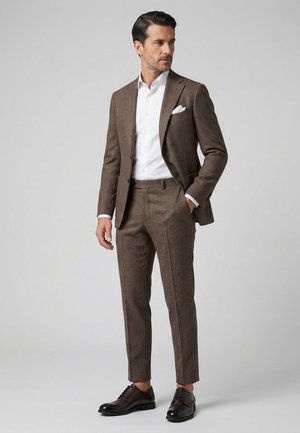 Homme debout portant un costume marron texturé, une chemise blanche, un mouchoir blanc dans la poche et des chaussures marron foncé, regardant sur le côté.