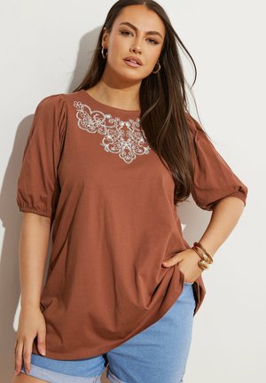 Vrouw met lang, donker haar, gekleed in een roestkleurige blouse met witte bloemrijke borduursels en lichtblauwe spijkerbroek, geaccentueerd met gouden armbanden en oorbellen.