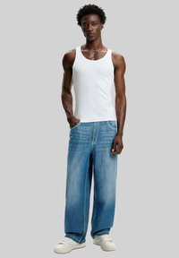 Man med kort lockigt hår som bär en vit tanktop, vida blå jeans och vita sneakers, stående med en hand i fickan.