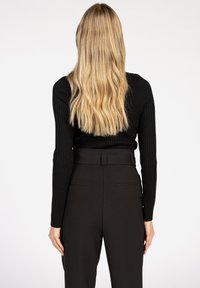 Vrouw met lang golvend blond haar, gekleed in een zwarte geribbelde trui en hoge zwarte broeken, staand met haar rug naar de camera tegen een effen achtergrond.