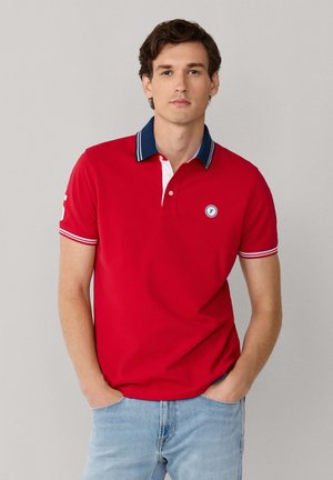 Joven con cabello castaño que lleva una camiseta tipo polo roja con cuello azul marino y jeans azul claro, de pie con las manos en los bolsillos.