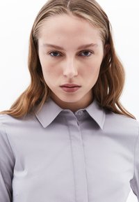Camisa de botón gris claro con cuello, textura suave y sutiles detalles de costura; el tejido parece resistente a las arrugas.