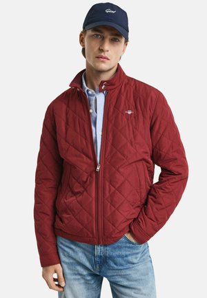 Red quilted jacket with a stand-up collar and zip closure, paired with a light blue shirt, and worn with blue jeans.
