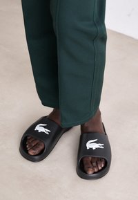 Des sandales noires à semelle ouverte, arborant un logo crocodile blanc. La personne porte un pantalon vert fuselé, mettant en valeur ses orteils.